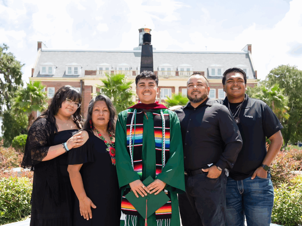 Luis Arriaga celebrates becoming the first in his family to obtain a four-year degree, surrounded by his parents and siblings. They are seen in front of the University's eternal flame, a moment symbolic of the motto, "Excellence With Caring."
